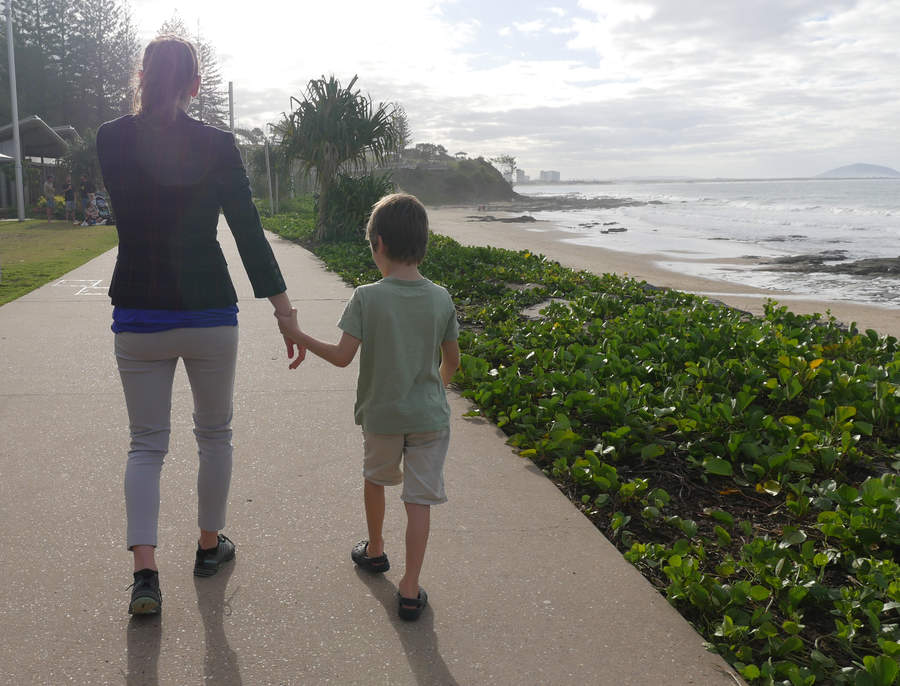 Woman providing sighted guide to young child holding her wrist, walking on a footpath near the beach.